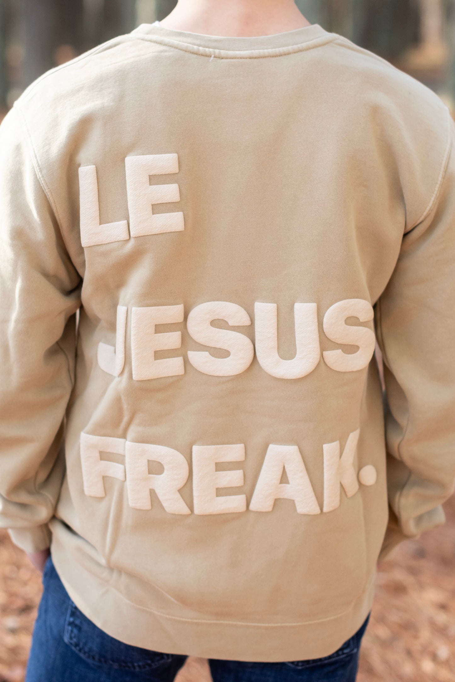 Beige sweatshirt with 'LE JESUS FREAK.' text on the back, worn by a person outdoors.